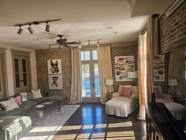 A spacious living room with a brick wall, a ceiling fan, and a large window with curtains. - Enjoy Island life in the Village in this gorgeous one of a kind loft! (St Simons Island)