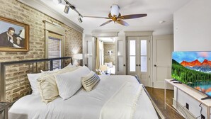 Un dormitorio con una cama grande, ventilador de techo, televisor montado y pared de ladrillo.