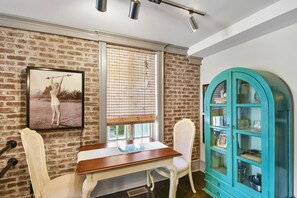 A dining area with a brick wall, a wooden table, two white chairs, a window with blinds, and a teal cabinet. - Enjoy Island life in the Village in this gorgeous one of a kind loft! (St Simons Island)