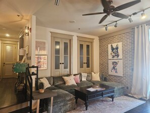 A living room with a sectional sofa, a coffee table, a ceiling fan, and artwork on the wall. - Enjoy Island life in the Village in this gorgeous one of a kind loft! (St Simons Island)