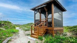 A wooden beach hut with a staircase leading up to a deck, surrounded by sand and vegetation. - Classic Oceanfront beach home with large screened in porch overlooking ocean! (Isle of Palms)