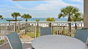 A balcony with a white table and chairs, overlooking a beach with palm trees and a wooden deck. - Sandy Key 231:  3 BR / 2 BA Gulf Front Condo in Perdido Key, FL  Sleeps 8 (Pensacola)