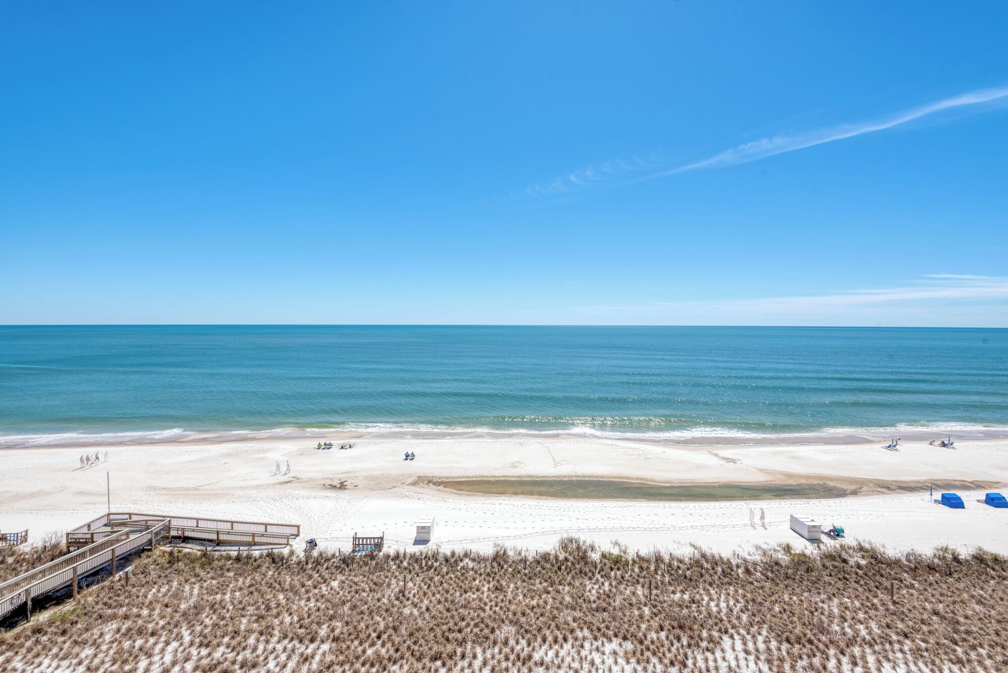 A beach with a wooden boardwalk leading to a sandy area, a clear blue sky, and calm ocean waters.