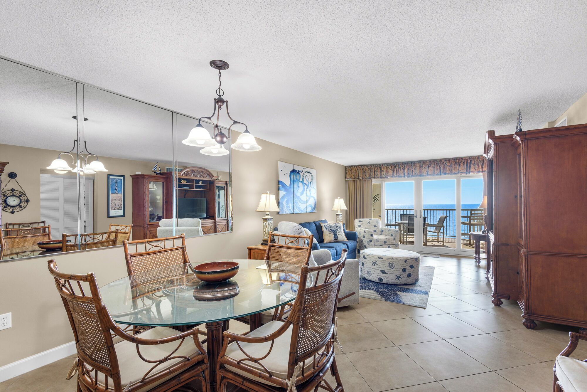 A spacious living area with a glass dining table surrounded by wicker chairs, a large mirror, and a view of the ocean through a sliding glass door.