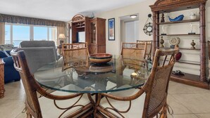 Un comedor con mesa de vidrio rodeada de sillas de mimbre, un aparador de madera con objetos decorativos y vistas al exterior a través de amplios ventanales.