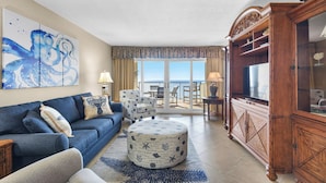 Un salón con sofá, reposapiés y sillón, un mueble de madera y un ventanal con vistas al mar.