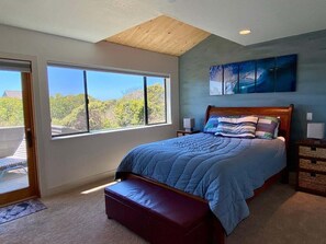 A bedroom with a large window, a bed with a blue bedspread, a wooden headboard, and a painting on the wall.