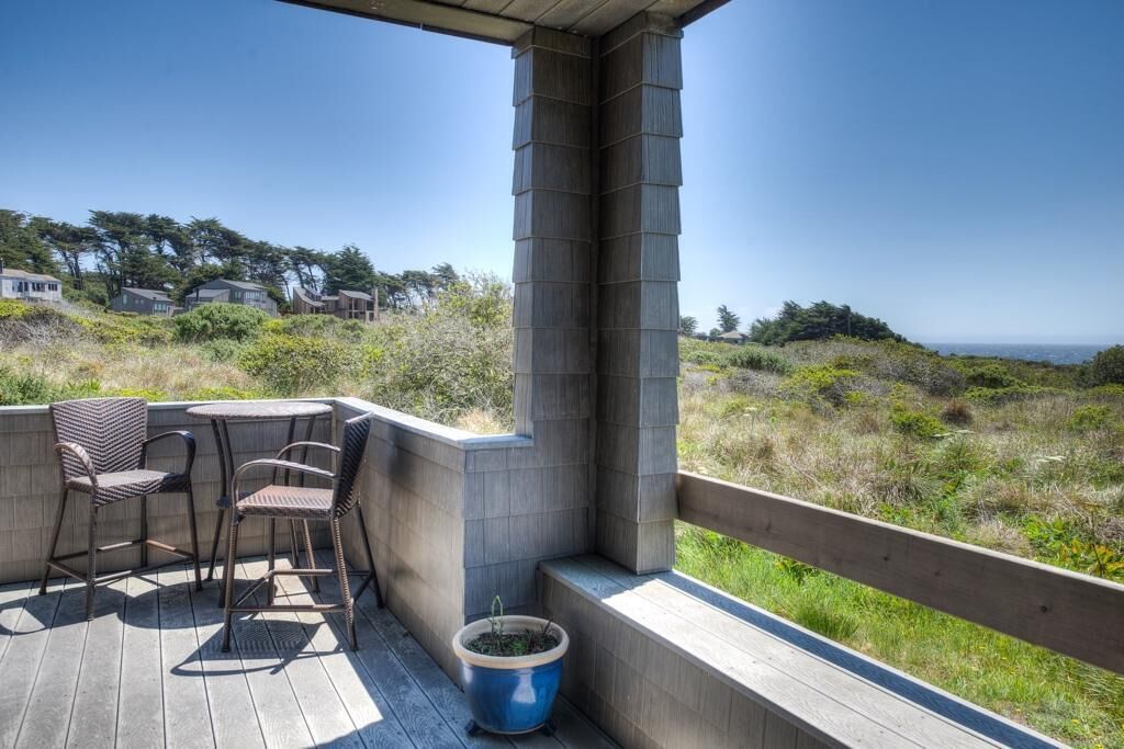 Eine Holzterrasse mit Gartenmöbeln, einem Blumentopf und Blick auf das Meer und Häuser im Hintergrund.