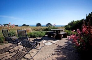 An outdoor seating area with chairs and a table, surrounded by a garden with flowers and a view of the ocean. - Brown-Hansen - Whitewater Sea Ranch Escape (Sea Ranch)