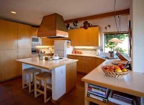 A kitchen with wooden cabinets, a central island with stools, and a fruit basket on the counter. - Brown-Hansen - Whitewater Sea Ranch Escape (Sea Ranch)