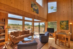 A spacious living room with a large window, a sofa, a television, and a dining area. - Clisham - Sea Ranch Cozy 2BR Loft Home (Sea Ranch)