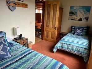 A room with two beds, a wooden door, a painting, and a small table with a lamp.