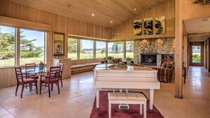 A spacious room with a piano, dining table, and chairs, large windows, and a stone fireplace. - Wild Iris | Ocean View Haven with Hot Tub (Sea Ranch)