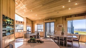 A modern kitchen with wooden cabinetry, a central island, and a dining area with a view. - Wild Iris | Ocean View Haven with Hot Tub (Sea Ranch)