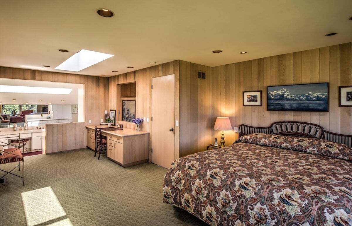 A bedroom with a large bed, a desk, and a chair. There is a skylight above the bed and a painting on the wall.