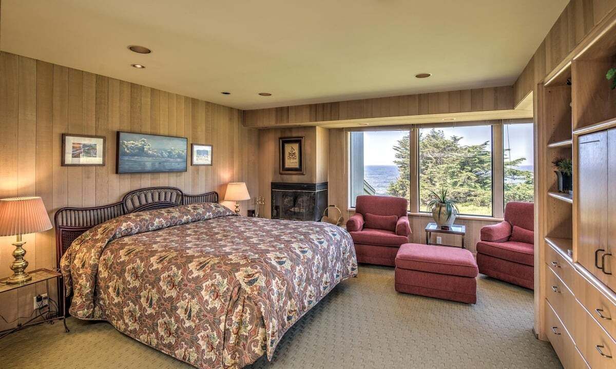 A bedroom with a large bed, a fireplace, a red armchair, and a view of the outdoors.