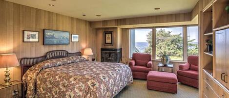 A bedroom with a large bed, a fireplace, a red armchair, and a view of the outdoors.