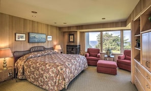 A bedroom with a large bed, a fireplace, a red armchair, and a view of the outdoors. - Wild Iris | Ocean View Haven with Hot Tub (Sea Ranch)