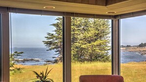 A room with large windows offering a view of the ocean and a forest. There are two red chairs and a vase with a plant on a table. - Wild Iris | Ocean View Haven with Hot Tub (Sea Ranch)