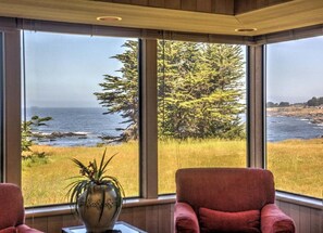 Ein Zimmer mit großen Fenstern, das einen Blick auf den Ozean und einen Wald bietet. Zwei rote Sessel und eine Tischvase mit Pflanze sind vorhanden.