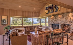 A spacious living room with a stone fireplace, large windows offering a view of the outdoors, and a high ceiling with wooden paneling.