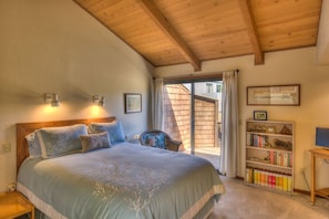 A bedroom with a large bed, wooden ceiling beams, a bookshelf, and a window with curtains.