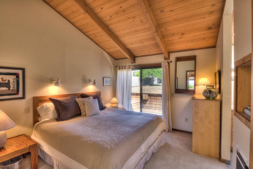 A bedroom with a large bed, wooden ceiling beams, a window with curtains, and a bedside table with a lamp.