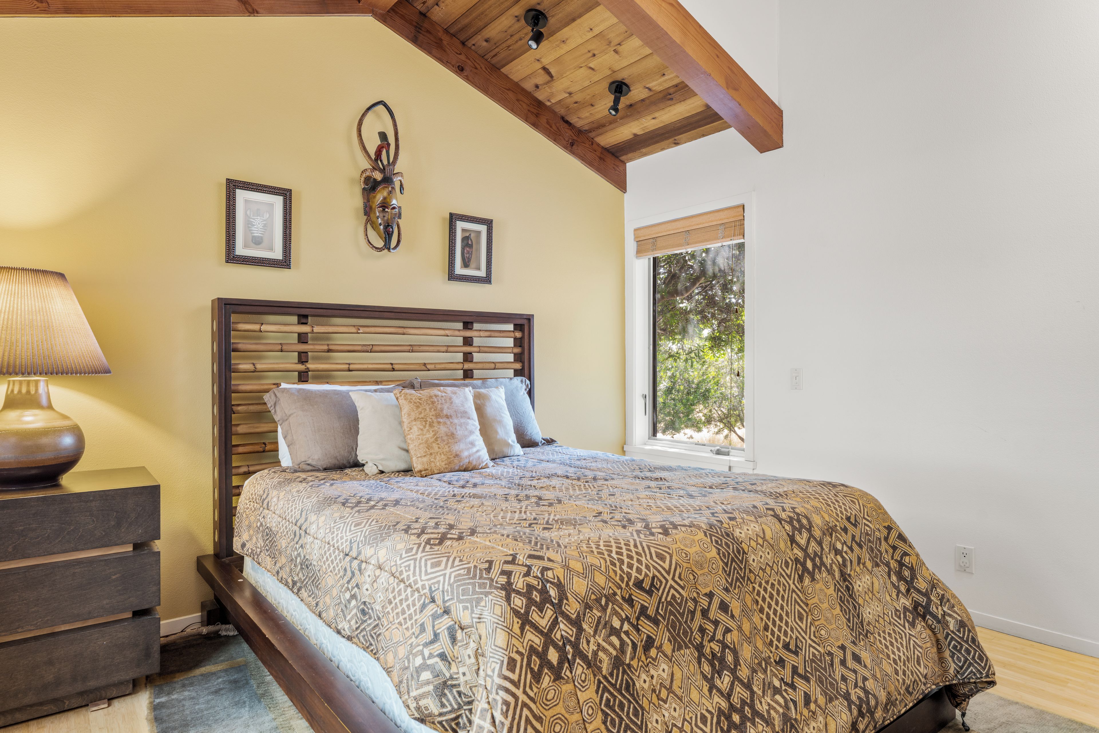 A bedroom with a wooden headboard, a patterned bedspread, a nightstand with a lamp, and two framed pictures on the wall.
