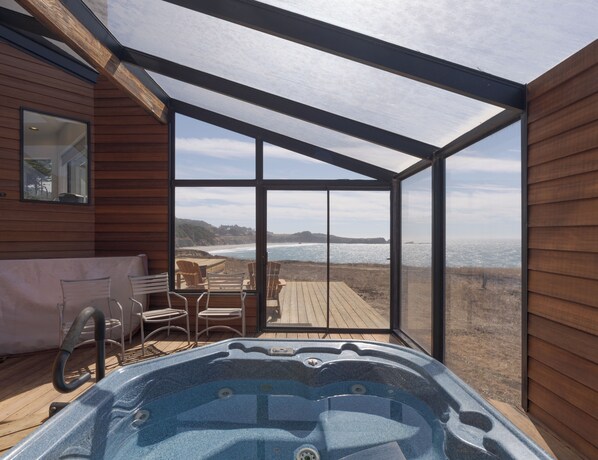 A hot tub on a wooden deck with a glass enclosure overlooking a beach.