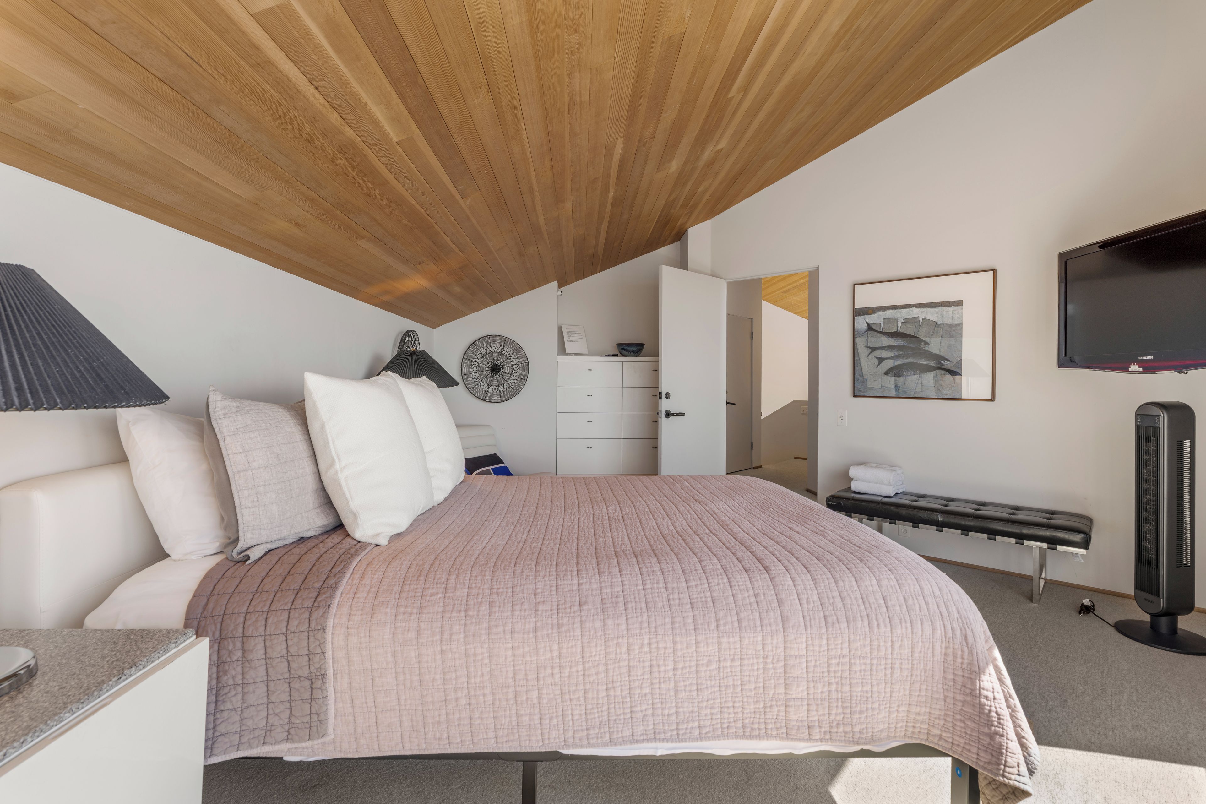 A bedroom with a sloped ceiling, a bed with a pink quilt, a bedside table, a lamp, a dresser, a mirror, a framed picture, a bench, and a heater.