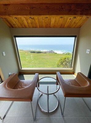 A room with a large window overlooking the ocean, two brown chairs, and a round table. - Sea Walk Retreat | Front-Row Whale Watching Condo (Sea Ranch)