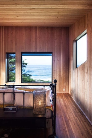 Un dormitorio con cama, mesita de noche, lámpara y vistas al océano a través de amplias ventanas.