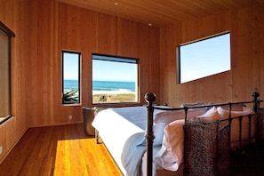 A bedroom with a wooden floor, a bed with a metal frame, two windows with a beach view, and a large window on the wall.