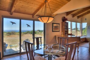 A dining area with a glass table and chairs, a wooden ceiling, and large windows with a view of the outdoors.