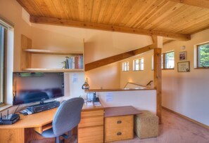 A room with a wooden desk, a television, and a chair. There are bookshelves above the desk and a window with a view of greenery.