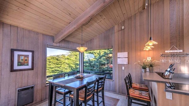 A wooden interior with a dining area, kitchen, and large windows overlooking greenery.