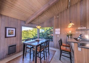 A wooden interior with a dining area, kitchen, and large windows overlooking greenery.