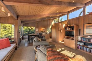 A spacious living area with a wooden ceiling, large windows, a sofa, a television, and a bookshelf.