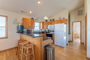 A kitchen with wooden cabinets, a central island with stools, a stainless steel trash can, and a refrigerator.