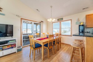 A dining area with a wooden table and chairs, a television on a white cabinet, and a kitchen counter with stools.