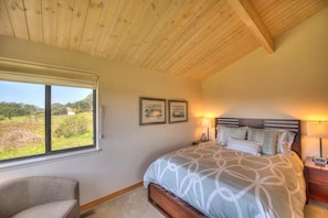 A bedroom with a wooden ceiling, a bed with patterned bedding, a nightstand with a lamp, and a window with a view of greenery.