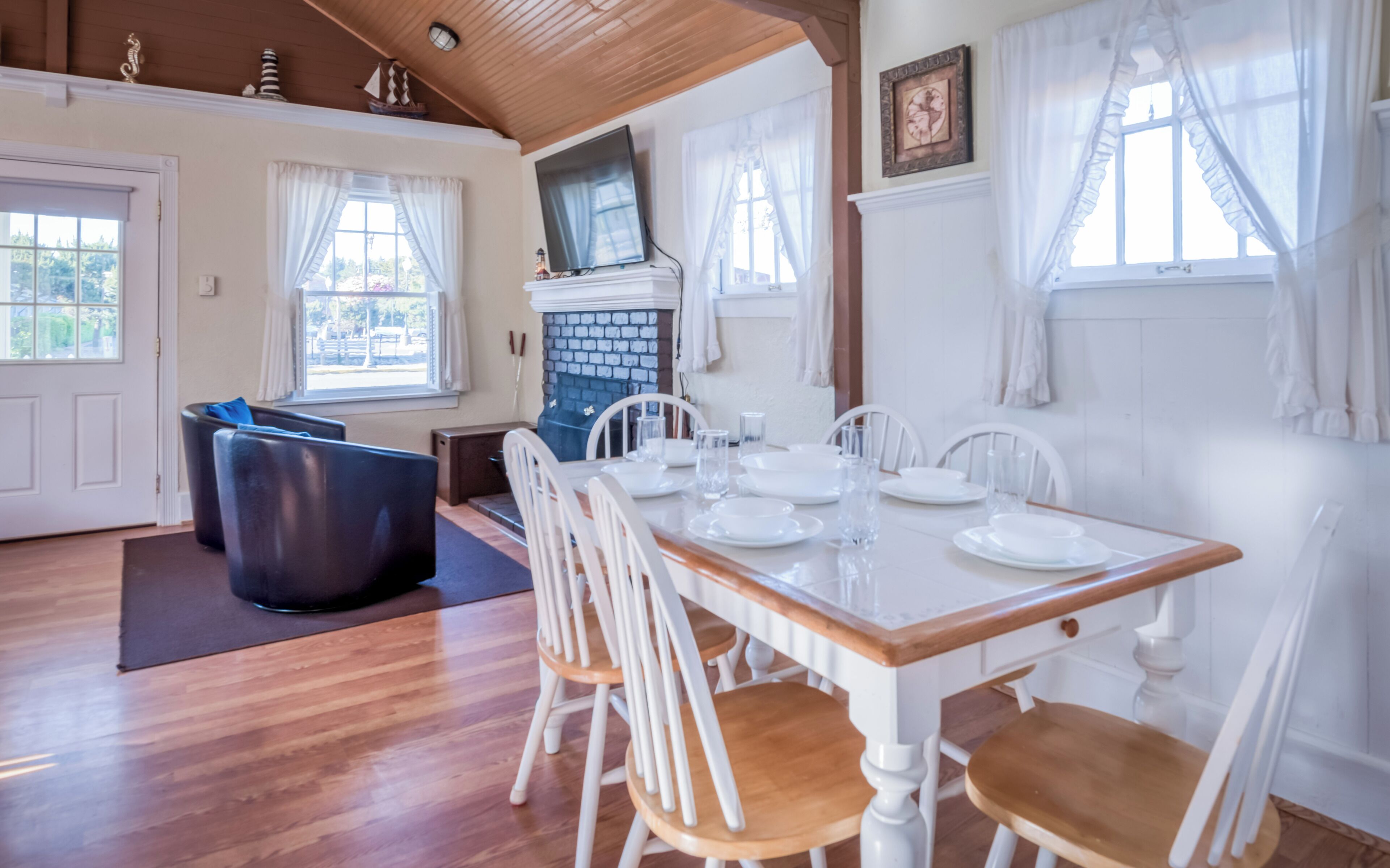 Un coin repas avec une table en bois dressée pour six personnes, des chaises blanches et une cheminée en briques.