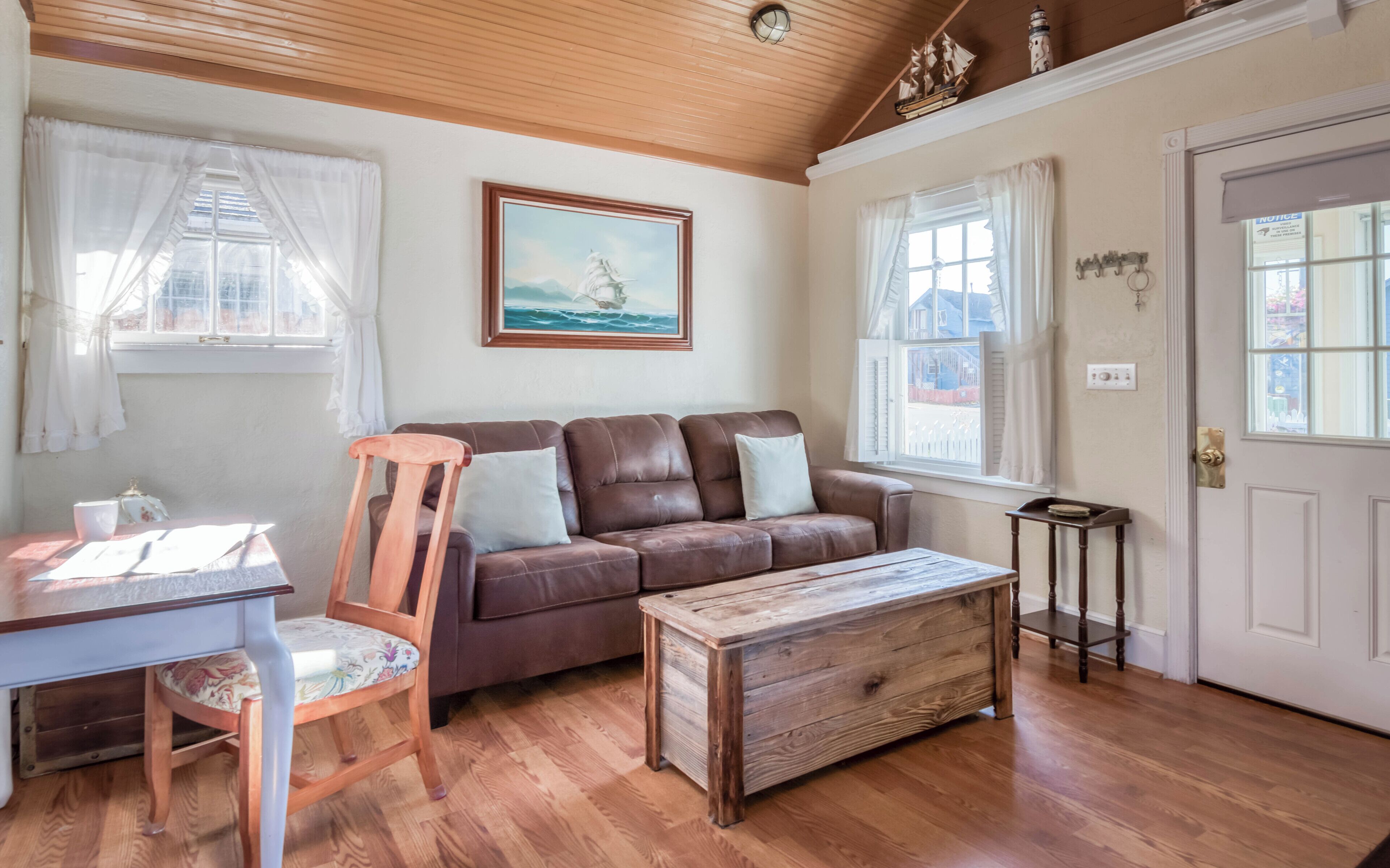 Una habitación acogedora con un sofá de cuero marrón, una mesa de centro de madera y una silla de madera. Hay un cuadro enmarcado en la pared, una ventana con cortinas y una puerta con un cartel.
