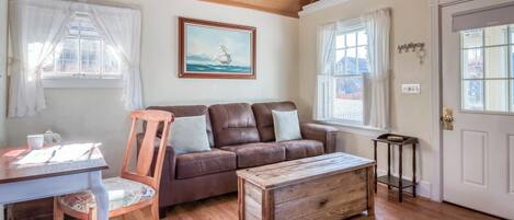 Una habitación acogedora con un sofá de cuero marrón, una mesa de centro de madera y una silla de madera. Hay un cuadro enmarcado en la pared, una ventana con cortinas y una puerta con un cartel.