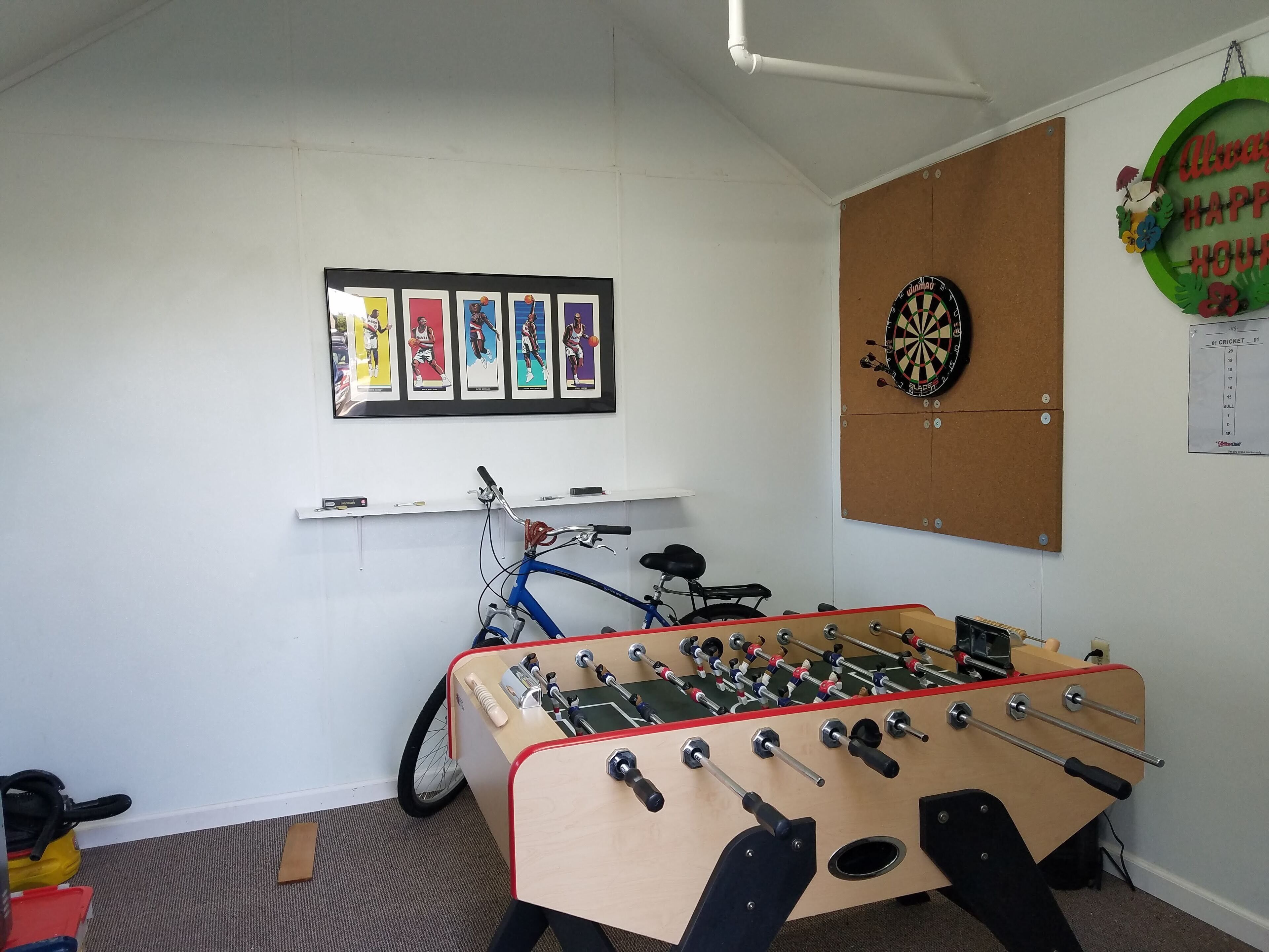 A room with a foosball table, a dartboard, and framed sports posters on the wall.