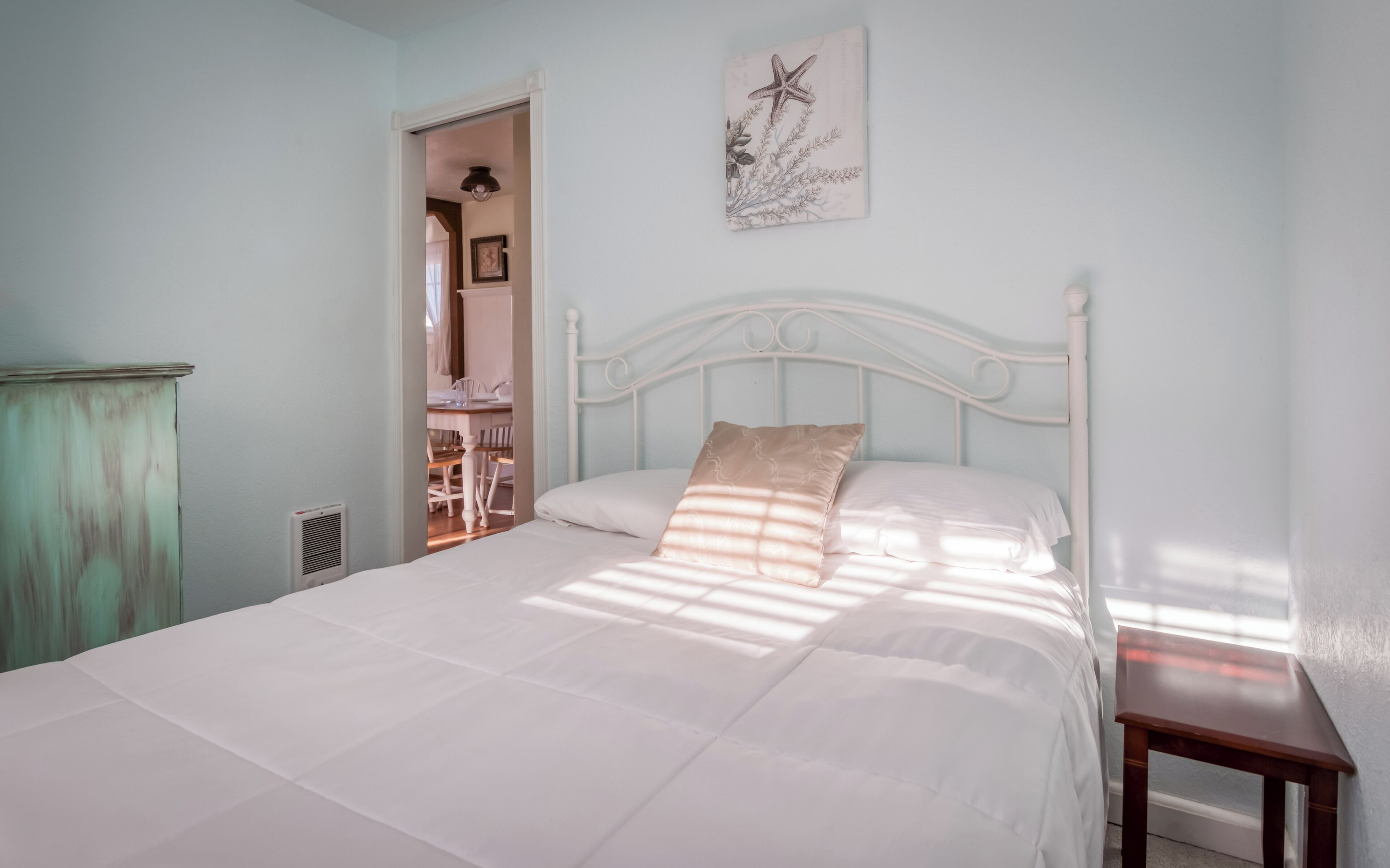 Un dormitorio con una cama blanca, una mesita de noche de madera y un cuadro enmarcado en la pared, además de una puerta que da a otra habitación.