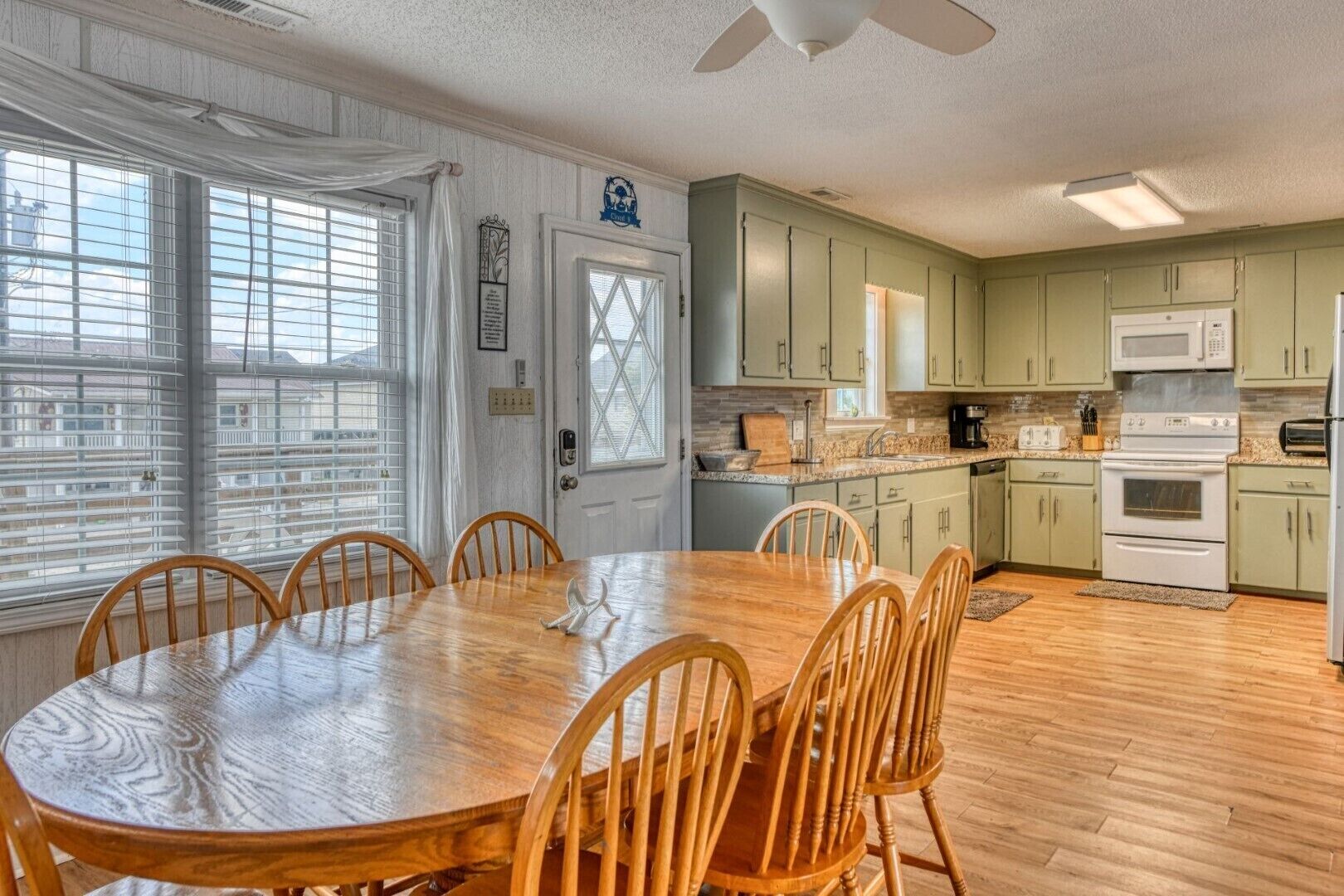 A modern kitchen with green cabinets, a wooden dining table, and a large window with blinds.