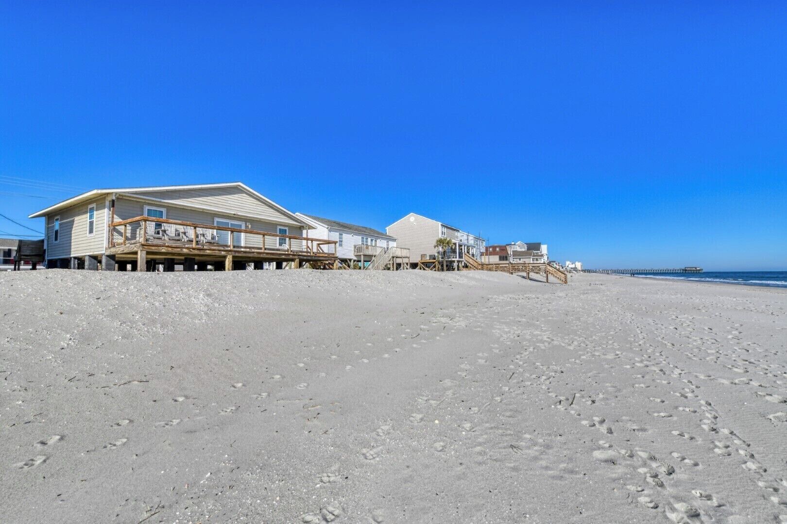 A beachfront property with multiple houses connected by wooden walkways, a pier extending into the ocean, and a clear blue sky.