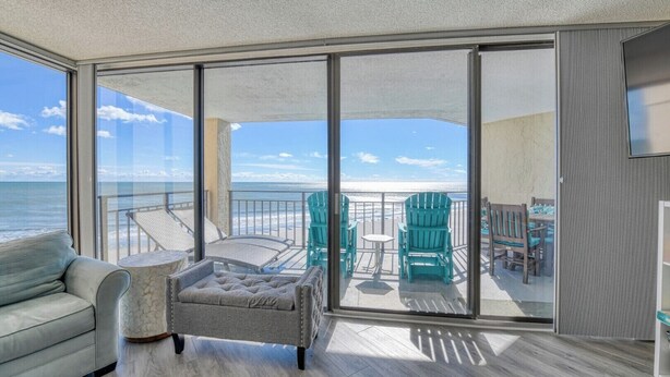 En balkon med udsigt over stranden, en grå sofa og en polstret lænestol.