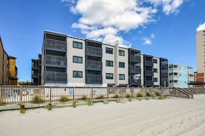 A multi-story residential building with balconies, a sandy beach in the foreground, and a clear blue sky.